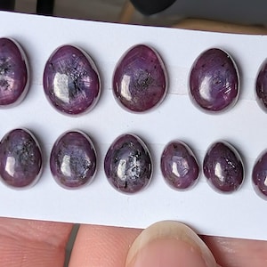 May include: A collection of thirteen oval-shaped, cabochon-cut star rubies arranged in two rows on a white display card. The gemstones exhibit a deep, dark reddish-purple hue with visible internal inclusions and a smooth, polished surface finish.