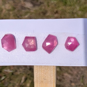 Op de afbeelding: Vier gefacetteerde, roze edelstenen worden getoond op een witte kaart, bevestigd aan een houten stok. De edelstenen hebben onregelmatige vormen en reflecteren licht. De achtergrond is wazig.