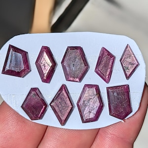 Op de afbeelding: Een verzameling van negen gefacetteerde, rood-paarse edelstenen in verschillende geometrische vormen, waaronder diamanten en zeshoeken. De edelstenen worden gepresenteerd op een wit oppervlak, waarbij hun gepolijste oppervlakken en interne patronen worden getoond.