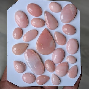May include: A collection of polished, oval, and teardrop-shaped pink gemstones arranged on a white surface. The gemstones vary in size and shade, with a soft, glossy appearance. The overall color palette is a delicate, pastel pink.