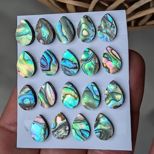 Puede incluir: Una colección de veinte cabujones en forma de lágrima de concha de abulón. Las conchas iridiscentes muestran una gama de colores, incluyendo azules, verdes y púrpuras, con patrones únicos. Los cabujones están dispuestos sobre una tarjeta gris.