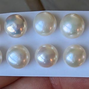 Könnte beinhalten: Zehn glänzende, runde Perlen in cremeweißer Farbe sind auf einer weißen Karte angeordnet. Jede Perle reflektiert schillernde Farben, darunter Rosa, Blau und Grün, wodurch ein schimmernder Effekt entsteht. Die Perlen sind ein klassischer Schmuckbestandteil.