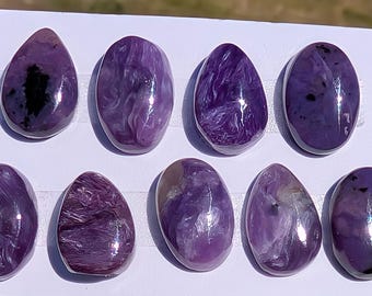 May include: Ten polished, oval charoite gemstones. The stones are various shades of purple, with swirling patterns and dark inclusions. The gemstones are displayed on a white surface, highlighting their natural beauty and unique textures.