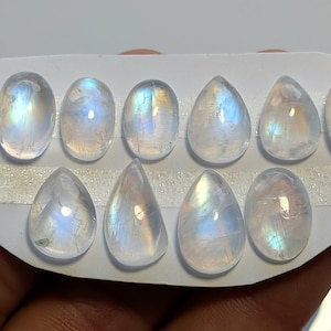 May include: Eleven iridescent moonstone gemstones in assorted shapes, including oval and teardrop, are displayed on a white, textured surface. The stones exhibit milky white and blue colours. The gemstones are held by a hand.