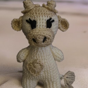 May include: A handmade, knitted cow figurine in cream and tan. The cow has black eyes, a tan muzzle, ears, and horns, and a heart-shaped patch on its chest. The tail is a small, gray tuft.