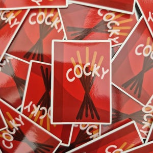 Puede incluir: Una colección de pegatinas rojas con la palabra "COCKY" en letras blancas. Las pegatinas presentan un gráfico de palitos de galleta cubiertos de chocolate. El fondo es una mezcla de tonos rojos.