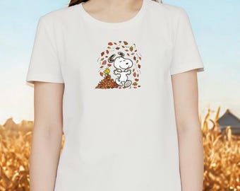 May include: White t-shirt featuring a cartoon illustration of Snoopy and Woodstock playing in autumn leaves. The graphic shows Snoopy standing next to a pile of leaves with Woodstock nearby, surrounded by falling leaves.