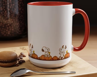 May include: White ceramic mug with a red interior and handle. The mug features a cartoon design of Snoopy and other Peanuts characters with pumpkins. A spoon and a macaron are on a wooden surface.