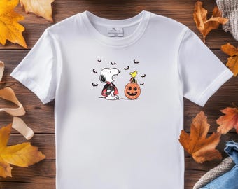 May include: White t-shirt featuring Snoopy in a vampire costume with Woodstock and a jack-o'-lantern. The design includes black bats and is set against a wooden background with autumn leaves, perfect for Halloween.
