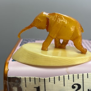 May include: A translucent amber-colored elephant figurine standing on a smooth, cream-colored oval base. A white measuring tape is positioned in the foreground, showing the base has a width of approximately 2.25 inches from the left edge.
