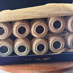May include: A blue cardboard box containing ten balls of beige DMC size 30 crochet thread. Each ball features a black circular label with the text DMC 30. The box is partially covered by a piece of light brown tissue paper tucked inside the top.