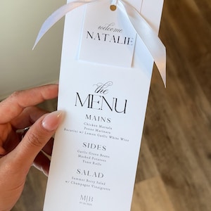May include: A white menu card with a ribbon, featuring the text "welcome NATALIE" at the top. The menu lists mains, sides, and salad options, including Chicken Marsala and Summer Berry Salad. The card is held by a person.