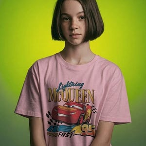 Puede incluir: Una camiseta rosa con un dise&ntilde;o de Rayo McQueen de la pel&iacute;cula Cars. El gr&aacute;fico incluye el texto "Lightning McQueen", "95" y "Turn Fast". La camiseta la lleva una persona con pelo corto casta&ntilde;o sobre un fondo verde.