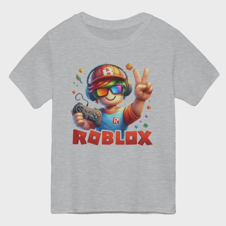 May include: A gray t-shirt featuring a colorful Roblox character with headphones, rainbow glasses, and a game controller. The character is giving a peace sign. The word "ROBLOX" is printed in red block letters.