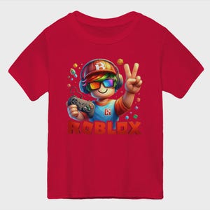 May include: Red t-shirt featuring a colorful graphic of a Roblox character wearing headphones and sunglasses, holding a game controller, and making a peace sign. The word "ROBLOX" is displayed in large, 3D letters below the character.