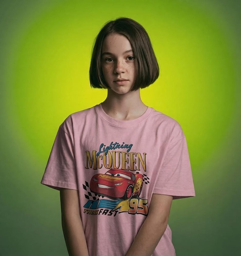 Puede incluir: Una camiseta rosa con un dise&ntilde;o de Rayo McQueen de la pel&iacute;cula Cars. El gr&aacute;fico incluye el texto "Lightning McQueen", "95" y "Turn Fast". La camiseta la lleva una persona con pelo corto casta&ntilde;o sobre un fondo verde.
