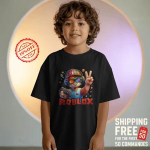 May include: Black t-shirt featuring a colorful Roblox character with rainbow sunglasses, a red and blue hat, and a controller. The word "ROBLOX" is printed in red. The shirt is worn by a child.