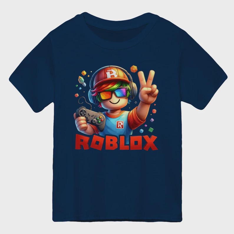 May include: Navy blue t-shirt featuring a cartoon gamer character with rainbow-colored glasses, headphones, and a controller. The word "ROBLOX" is printed in red block letters below the character. The character is giving a peace sign.