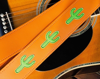 Peut inclure: Une sangle de guitare orange avec des motifs de cactus verts. La sangle est posée sur une guitare acoustique.