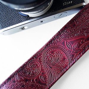 May include: A red leather camera strap with a paisley pattern. The strap is embossed with a detailed design.
