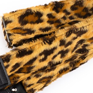 May include: A guitar strap featuring a faux fur leopard print pattern in shades of tan, brown, and black. The strap includes black leather ends with keyhole slots for attachment and a silver metal adjustment buckle. The text couch hand made is visible.