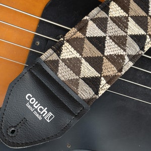 May include: A black and brown guitar strap with a geometric pattern. The strap is made of fabric and has a leather end with the text "couch" and "hand made".