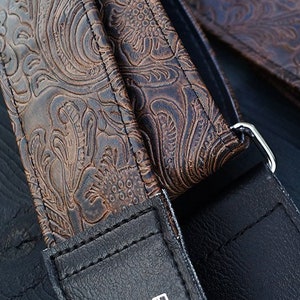 May include: Two brown leather guitar straps with an embossed floral pattern. The straps have a black leather backing and silver metal hardware.