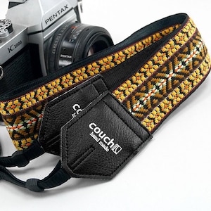 May include: A black camera strap with a yellow, orange, and brown geometric pattern. The strap has two black leather ends with the text "couch hand made" printed in white.