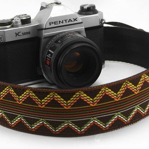 May include: A silver Pentax K1000 camera with a black lens and a brown camera strap. The strap features a repeating zigzag pattern in white, yellow, orange, and green on a brown background.