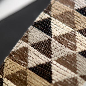 May include: A close-up of a guitar strap with a geometric pattern in shades of brown, beige, and black. The strap is made of a woven fabric with a textured surface.