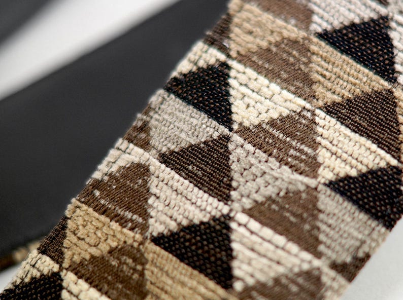May include: A close-up of a guitar strap with a geometric pattern in shades of brown, beige, and black. The strap is made of a woven fabric with a textured surface.
