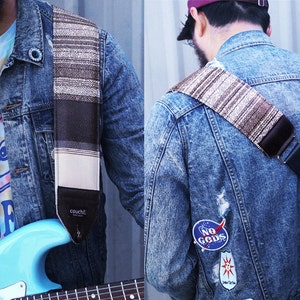 Puede incluir: Una persona lleva una chaqueta vaquera con parches y una camiseta de tie-dye y sostiene una guitarra el&eacute;ctrica azul con una correa de guitarra marr&oacute;n y blanca. La correa tiene la palabra "Couch" impresa.