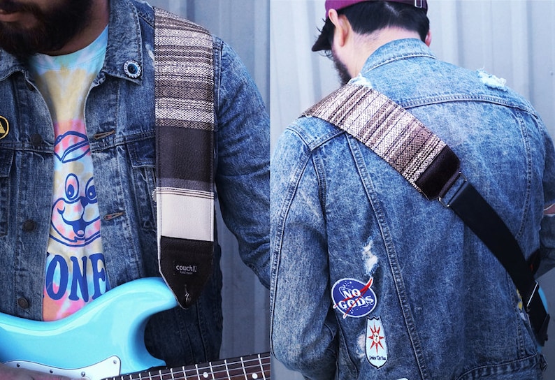 Puede incluir: Una persona lleva una chaqueta vaquera con parches y una camiseta de tie-dye y sostiene una guitarra el&eacute;ctrica azul con una correa de guitarra marr&oacute;n y blanca. La correa tiene la palabra "Couch" impresa.