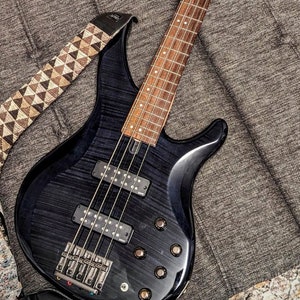 May include: A black electric bass guitar with a brown wooden fretboard and a patterned guitar strap. The guitar is lying on a gray textured surface.