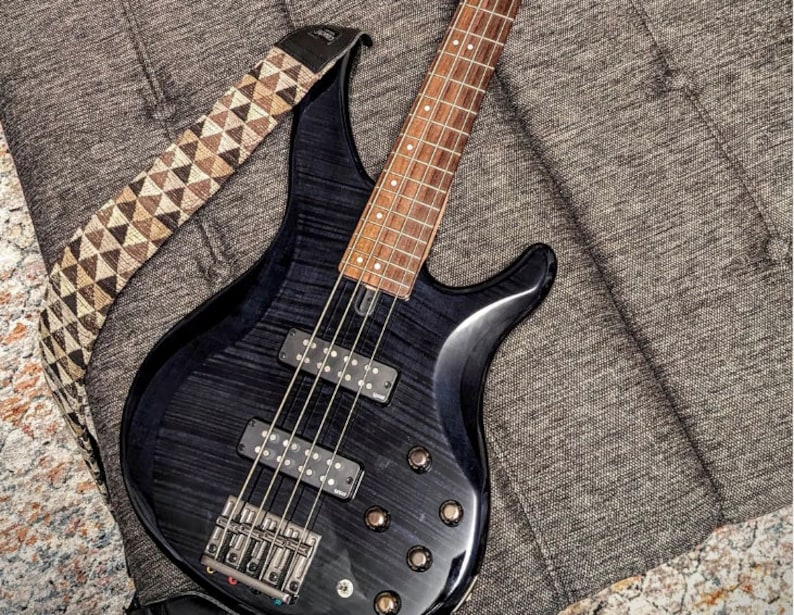 May include: A black electric bass guitar with a brown wooden fretboard and a patterned guitar strap. The guitar is lying on a gray textured surface.