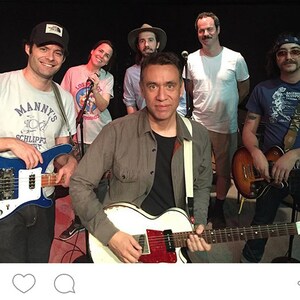 May include: A group of musicians, The Blue Jean Committee, are shown in a rehearsal setting. The band members are holding guitars and microphones. The image includes the text "Manny's" on a t-shirt and the hashtag #bluejeancommittee.