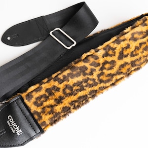 May include: A guitar strap with a black nylon strap and a leopard print section. The leopard print is brown and tan. The strap has a metal buckle and black leather ends with the brand name "couch band made".