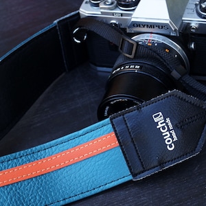 May include: A teal blue camera strap with an orange stripe and black leather accents. The strap has the text "couch" and "hand made" printed on it.