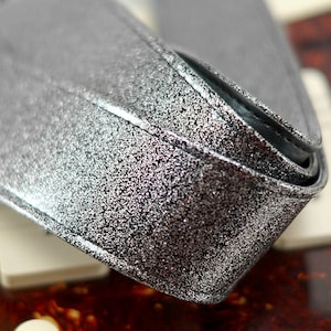 Pu&ograve; includere: Primo piano di una tracolla per chitarra argentata con glitter. La tracolla &egrave; realizzata in un materiale spesso e resistente e ha una finitura liscia e lucida.