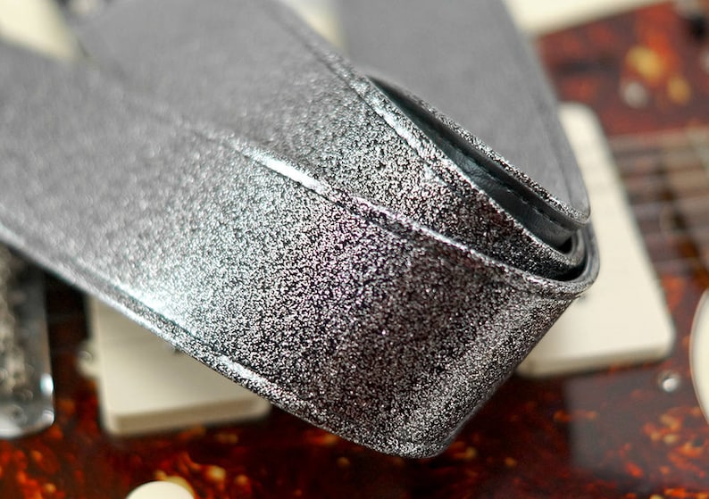 Pu&ograve; includere: Primo piano di una tracolla per chitarra argentata con glitter. La tracolla &egrave; realizzata in un materiale spesso e resistente e ha una finitura liscia e lucida.