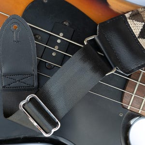 May include: A black and brown guitar strap with a geometric pattern. The strap is attached to a black bass guitar with a wooden fretboard.