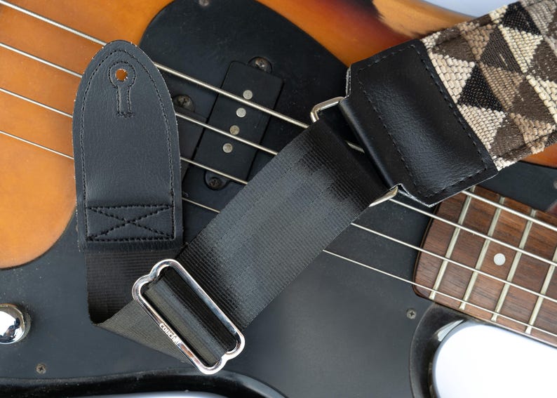 May include: A black and brown guitar strap with a geometric pattern. The strap is attached to a black bass guitar with a wooden fretboard.