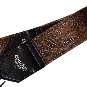 May include: Brown leather camera strap with an embossed floral pattern. The strap has a black leather patch with the text "couch" and "hand made" in white.