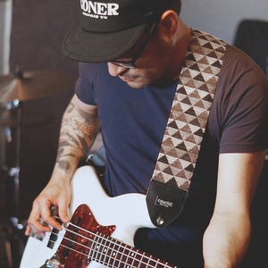 May include: A person playing a white bass guitar with a brown and white patterned guitar strap. The strap has the word "Couch" printed on it. The person is wearing a black t-shirt and a black baseball cap with the word "DONER" printed on it.