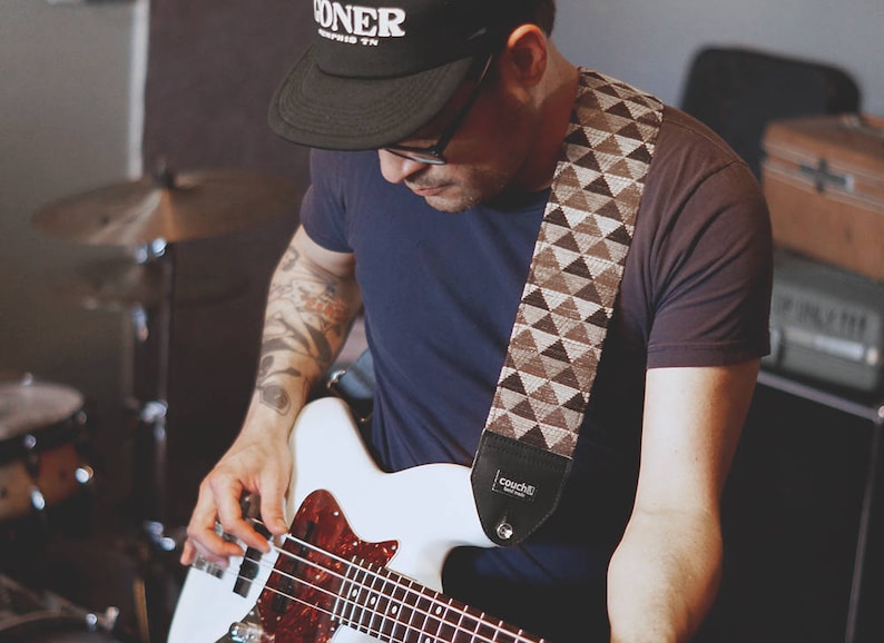 May include: A person playing a white bass guitar with a brown and white patterned guitar strap. The strap has the word "Couch" printed on it. The person is wearing a black t-shirt and a black baseball cap with the word "DONER" printed on it.