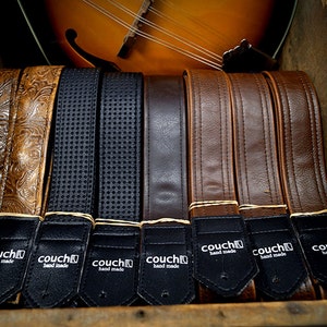 May include: A close-up of eight leather guitar straps in various colors and textures, including brown, black, and tan. The straps are arranged in a row and have a black label with the text "couch hand made" on each.