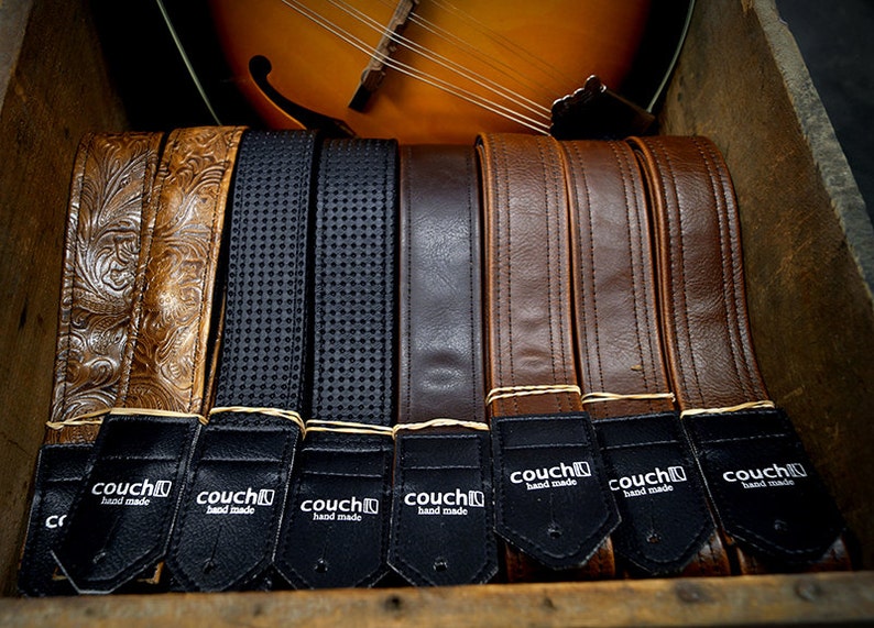 May include: A close-up of eight leather guitar straps in various colors and textures, including brown, black, and tan. The straps are arranged in a row and have a black label with the text "couch hand made" on each.