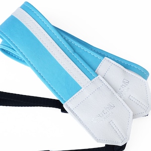 May include: A light blue camera strap with a white stripe and white leather ends. The leather ends have the text "Camera Strap" embossed on them.