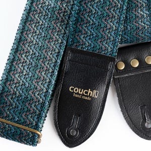 Könnte beinhalten: Ein gitarrengurt mit teal- und grauen Muster und schwarzen Lederenden. Der Gurt hat eine Metallschnalle und die Lederenden sind mit messingfarbenen Nieten befestigt. Der Text "couch hand made" ist sichtbar.