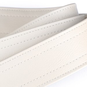 May include: Close-up of a cream-colored leather belt. The belt is folded over itself, showcasing the detailed stitching. The stitching is a lighter color than the belt, creating a subtle contrast. The leather appears smooth and supple.